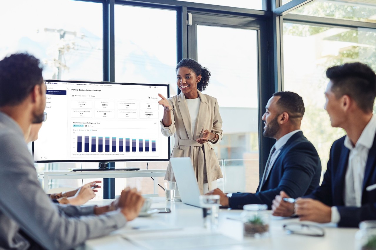 A business professional gives a presentation to colleagues in a modern conference room, using a large screen to display vehicle fleet emissions data.