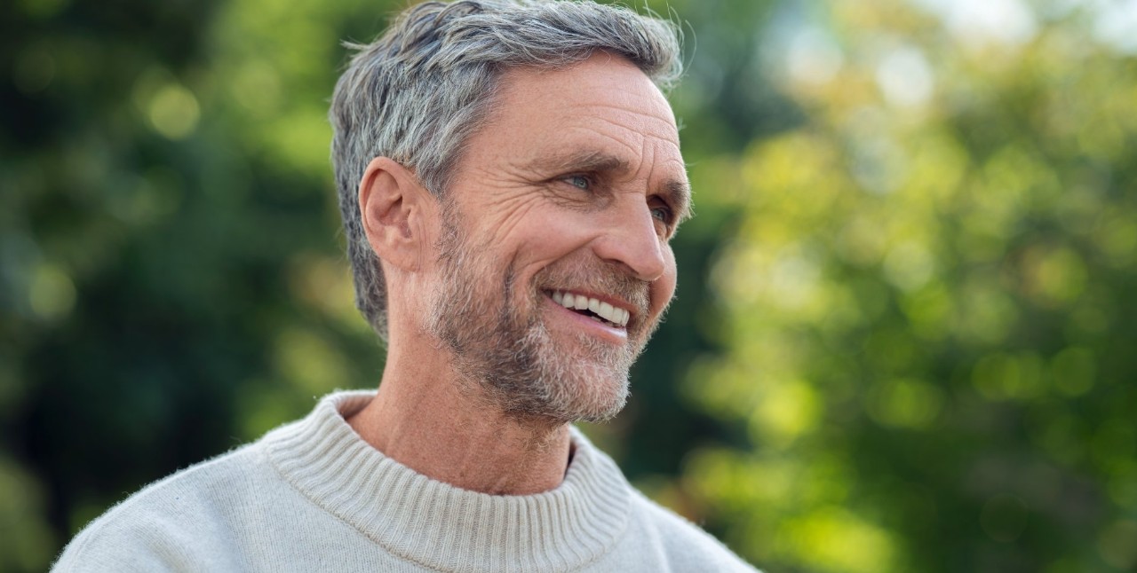 Man with gray hair and a light sweater stands outdoors and smiles.