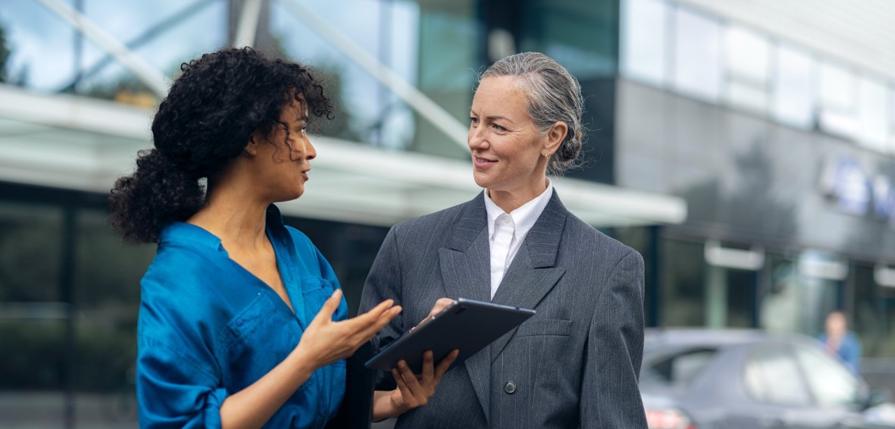 Two women have a conversation in front of a building with large glass windows.