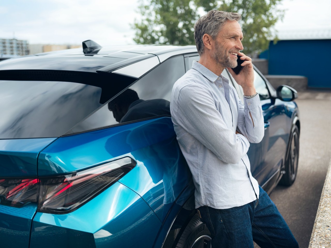 Man in a light shirt leans against a blue car while talking on a mobile phone in a parking lot.