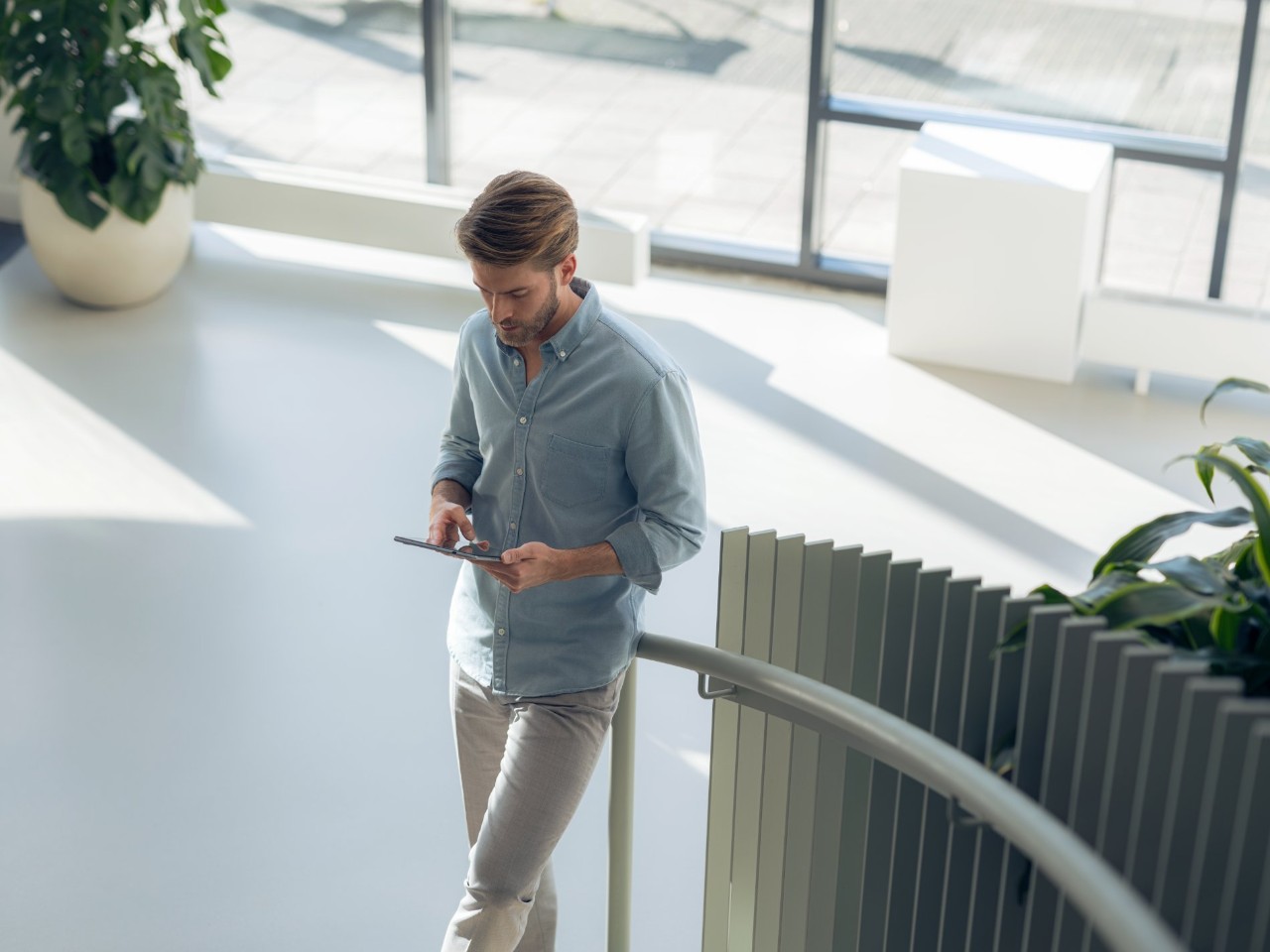 Man leans against a staircase railing with a tablet in a modern, well-lit room.