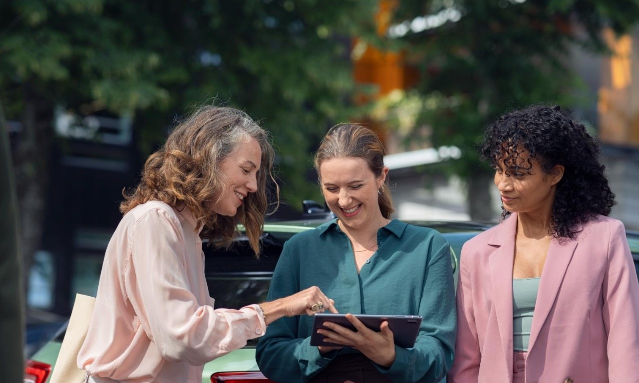 Three women discuss in front of a green car, holding a tablet.