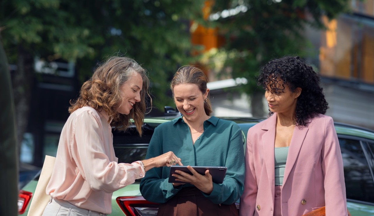Three women discuss in front of a green car, holding a tablet.